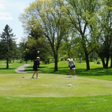 Two women tee off in front of trees